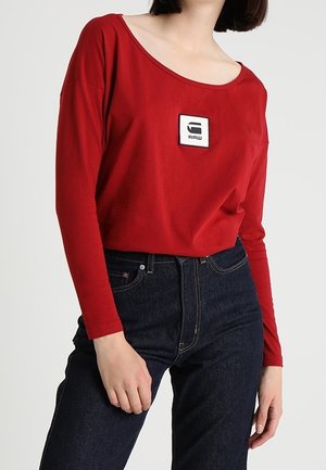 Kobieta w czerwonym długim rękawie z białą naszywką z logo "RAW" i ciemnoniebieskich jeansach z wysokim stanem na jasnym tle.