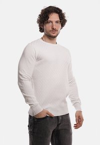 Maglione bianco strutturato con un motivo geometrico, polsini e orlo a coste, realizzato in un materiale knit morbido. Indossato con jeans scuri.