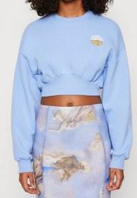 En person klädd i en ljusblå, kort sweatshirt med texten "Good Times" och en högmidjad kjol med ett tryck av en renässansstil cherub och moln.