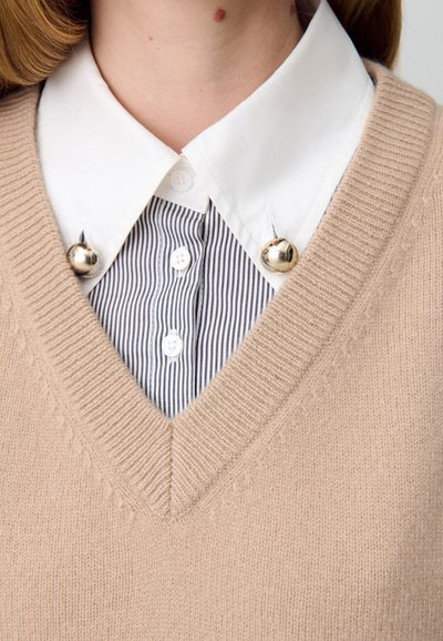Pull en tricot beige avec un décolleté en V superposé sur une chemise à col rayée blanche et marine, avec des accents en métal argenté au niveau du col.