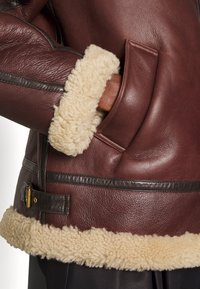 Gros plan sur une veste en cuir marron avec une doublure en peau lainée crème, montrant une main dans une poche latérale et un détail de ceinture à la taille.