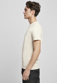 Beige T-shirt met korte mouwen van katoen met een ronde hals, gecombineerd met zwarte shorts, getoond in zijaanzicht tegen een effen achtergrond.