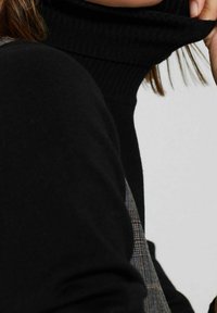 Zwarte coltrui met een geribbelde kraag, gecombineerd met een gestructureerde grijze, geprinte overlay. Focus op stof en ontwerpdDetails.