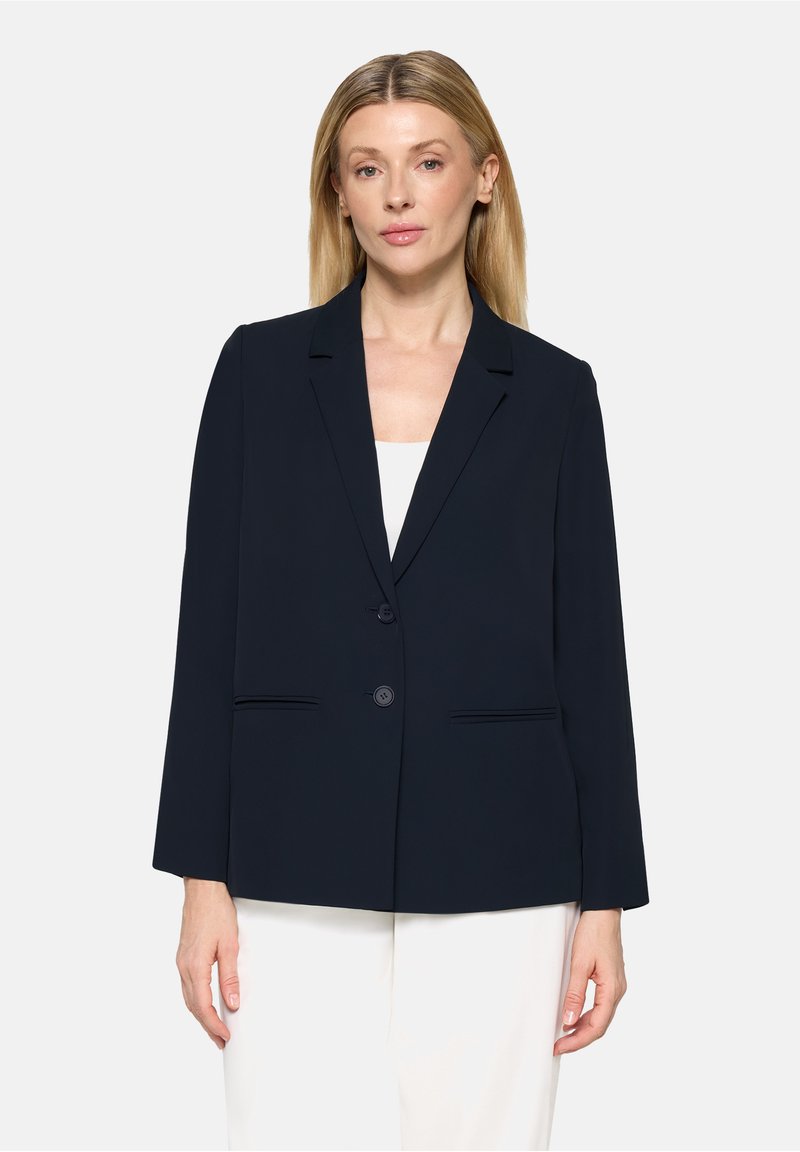 Femme aux cheveux blonds raides portant un blazer bleu marine à deux boutons sur un haut blanc et un pantalon blanc, faisant face à l’avant avec une expression neutre.