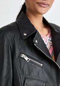 Veste en cuir noir avec une poche zippée, quincaillerie en argent, large col et finition texturée. Portée par-dessus un haut à motifs multicolores.
