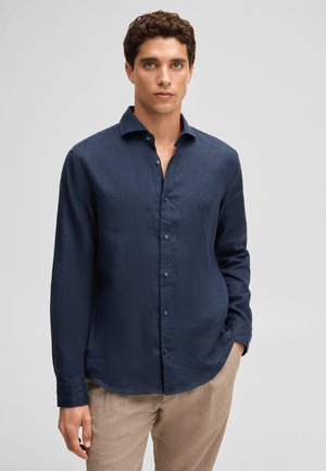 Mężczyzna ubrany w granatową, długorękawową koszulę z guzikami i beżowe spodnie, stoi z jedną ręką w kieszeni na jednolitym tle.