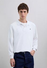 Polo blanc à manches longues avec un col et un petit emblème noir sur le côté gauche de la poitrine, associé à un jean denim bleu foncé.