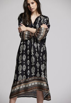 Schwarzes gemustertes Kleid mit geometrischen weißen Designs und einem braunen gemusterten Saum. Lange Ärmel und entspannte Passform, aus glattem Stoff gefertigt.