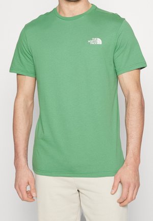 Mężczyzna ubrany w zielony T-shirt z krótkim rękawem z logo The North Face na piersi oraz beżowe spodnie na jednolitym tle.