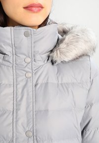 Harmaa pehmeä takki, jossa on korkea kaulus, napit ja pehmeä tekoturkki hupussa, joka tuo mukanaan sileän ja kiiltävän pinnan.