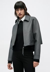 Osoba oblečená v sivom krátkom vetrovke na zips cez čierny sveter a bielu košeľu s golierom, stojaca pred jednoduchým bielym pozadím.