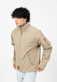 Beżowa kurtka z stójką, z przodu na zamek, z dwoma bocznymi kieszeniami oraz kieszenią na rękawie na zamek. Ozdobiona naszywkami.