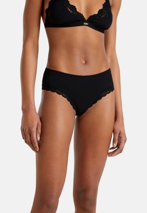 Donna che indossa un reggiseno nero con pizzo e culotte nere coordinate, mostrata dalla metà del petto fino a metà coscia su uno sfondo bianco.