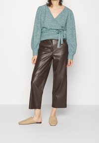 Turkos insvept tröja med långa ärmar och knytmidja, tillsammans med bruna läderbyxor med vida ben och vävt beigt slip-on skor.