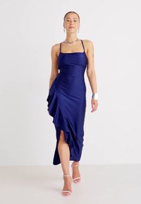 Navy blue satin dress with spaghetti straps, side draping, and a thigh-high slit. Model wearing clear heeled sandals and minimal accessories.