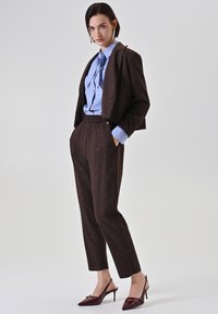 Femme debout, mains dans les poches, portant un costume marron foncé à fines rayures, un chemisier bleu clair avec un nœud, et des escarpins burgundy à bride arrière sur fond blanc.