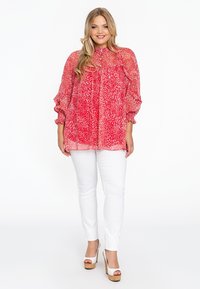 Rote Bluse mit weißem abstraktem Muster, hoher Kragen und Puffärmeln, kombiniert mit weißen Slim-Fit-Hosen und beigen Plateauschuhen.