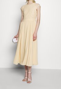 Kvinna i en ärmlös klänning i beige spets, som håller en vit clutch och bär vita remsandaler med hög klack mot en vit bakgrund.