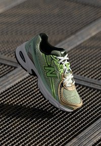 Lichtgroene en beige sneaker met bovenwerk van mesh, zwarte accenten, veters en een opvallend "N"-logo; heeft een dikke witte zool voor demping.