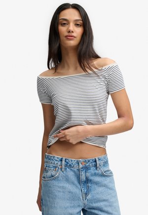 Jeune femme aux longs cheveux foncés portant un top rayé blanc et bleu marine dévoilant les épaules, et un jean taille haute bleu clair.
