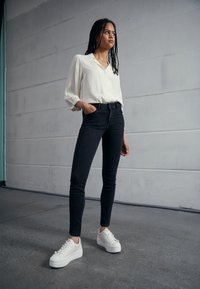 Ung kvinna med långt flätat hår står i en vit blus, svarta jeans och vita sneakers framför en enfärgad grå vägg.