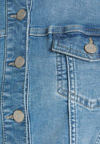 Džínová bunda z světlého modrého materiálu, se stříbrnými kovovými knoflíky, kapsou na hrudi a kontrastním prošíváním. Má hladkou texturu a strukturovaný střih.