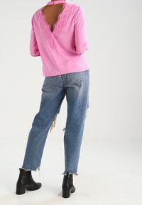 Blusa rosa a maniche lunghe con schiena aperta rifinita in pizzo, abbinata a jeans chiari strappati e stivaletti neri alla caviglia.