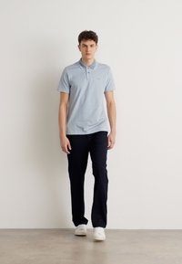 Lys blå polo shirt med tre knapper, kombinert med mørk marineblå bukser. Modellen har på seg hvite joggesko. Glatt stoff og skreddersydd passform.