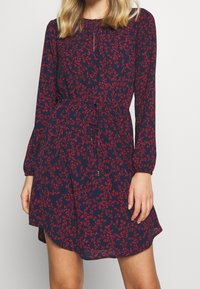 Robe bleu marine avec un imprimé floral rouge, manches longues, taille ajustable par un cordon, et une forme évasée décontractée en A, avec un col rond et une texture subtile.