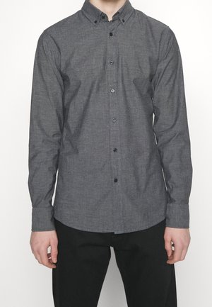 Mężczyzna ubrany w długą, grafitowo-szarą koszulę na guziki i czarne spodnie, stojący na jednolitym białym tle.