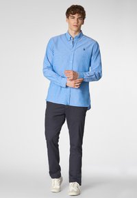 Camicia button-down azzurro chiaro con una sottile texture, caratterizzata da un piccolo logo sul petto, abbinata a pantaloni scuri e sneakers bianche.