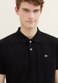 Zwarte polo met een kraag, twee knopen, klein logo op de borst, gemaakt van gestructureerde stof. Korte mouwen.