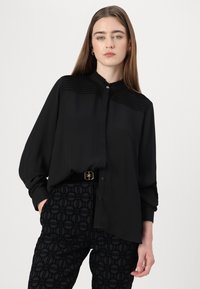 Zwarte blouse met geplooide details op de schouders, knoopsluiting aan de voorkant en losse mouwen, gecombineerd met zwarte Trousers met patroon en een gouden riem.