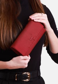 Red fabric wallet with a smooth texture, featuring an embroidered logo. Held in hand against a black outfit and light background.