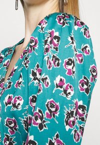 Türauf Stoffbluse in Blaugrün mit floralem Muster in Schwarz und Pink. Das Design umfasst einen V-Ausschnitt und dezente Drapierungen an den Ärmeln.