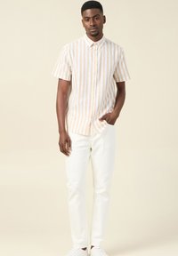 Camicia a maniche corte con bottoni, a strisce verticali beige chiaro e bianche. Indossata con jeans bianchi e sneakers bianche. Texture liscia.