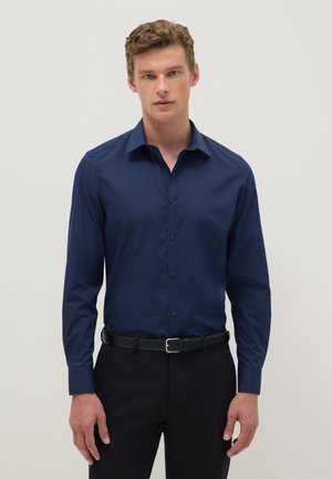 Hombre con camisa azul marino entallada abotonada y metida dentro de pantalones negros con cinturón negro, de pie contra un fondo liso.