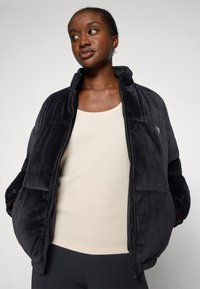 Giacca puffer nera in peluche con colletto alto, chiusura a zip e tasche laterali, indossata sopra un top a canotta rigato di colore beige chiaro, con una texture liscia.