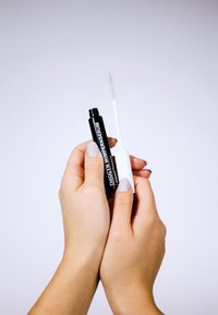 Black serum tube labeled "THIOCYAN WIMPERNSERUM" held in hands, paired with a white applicator wand with a fine, textured tip.