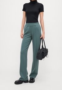 Svart ribbad polokrage med ljusgröna vida byxor med elastisk midja, hållande en svart handväska och kraftiga svarta skor.