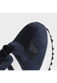 Marinblå sneaker med övre i mocka och mesh, med vita ränder, texturerade detaljer och en gummisula med ett robust mönster för bra grepp.