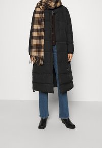 Person i lång svart pufferjacka, blå jeans, svarta ankelstövlar och en beige och brun rutig scarf på en ljus bakgrund.