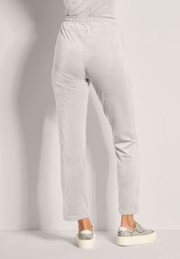 Persoon in lichtgrijze velours joggerbroek en witte platformsneakers met slangenprintdetails, staand tegen een vlakke muur.