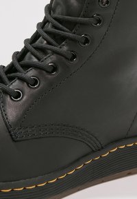 Bota de cuero negro con cordones, ojales de metal, costuras detalladas y una suela gruesa con costuras amarillas.