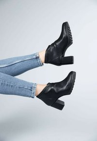 Black ankle boots made of leather, featuring a block heel and textured sole. Paired with blue skinny jeans, displayed against a light background.