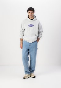 Sweatshirt à capuche gris avec logo violet, associé à un jean baggy bleu clair. Le modèle porte des baskets beiges et grises. Design simple et moderne.
