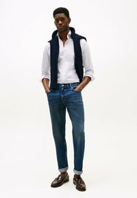 Camisa blanca de botones, suéter de punto de cable azul oscuro sobre los hombros, jeans de mezclilla azul, mocasines marrones, pose de pie. Fondo simple.