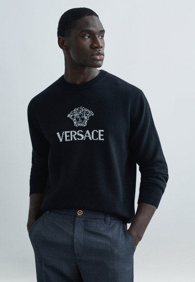 Black knitted sweater featuring a white "Versace" logo and graphic motif at the chest; worn with gray tailored trousers and a gold button detail.