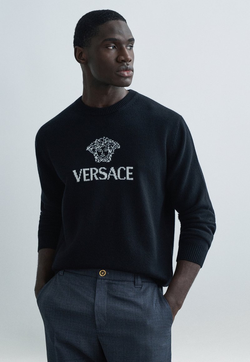 Černý pletený svetr s bílým logem "Versace" a grafickým motivem na hrudi; nošený se šedými na míru ušitými kalhotami a zlatým knoflíkovým detailem.