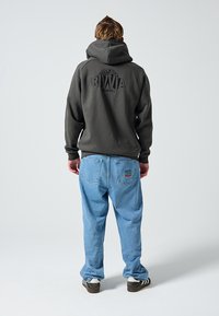 Tamnosivi hoodie s velikim crnim logom na leđima, uparen s svijetloplavim trapericama koje imaju šareni zakrpu na desnom džepu.